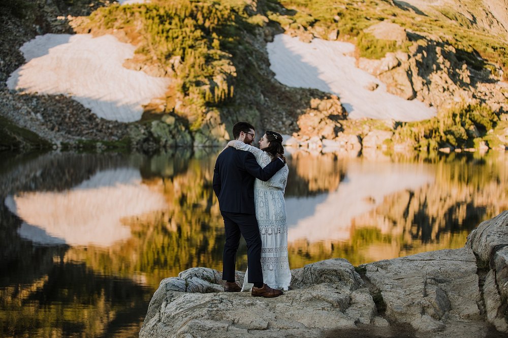 bride and groom standing together on alpine lake shore, calm glass like reflection on lake isabelle, patches of snow on summer mountains, colorado spring snow runoff, pawnee pass alpine lakes 