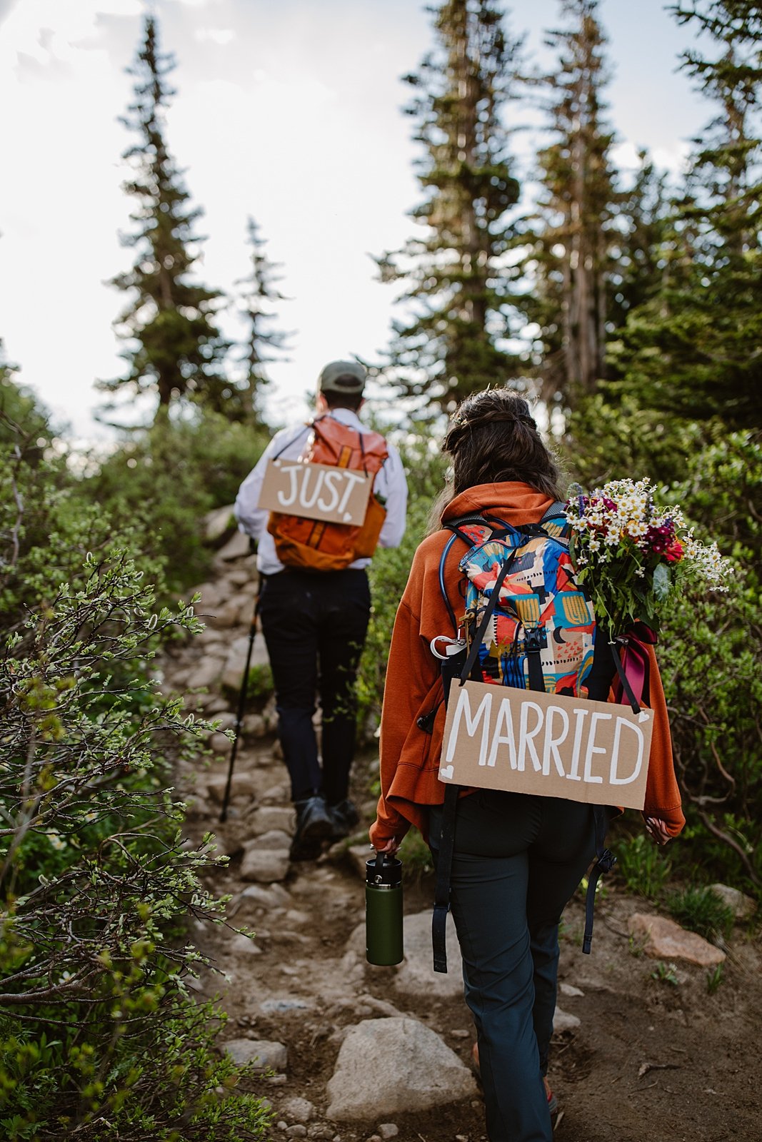 couple hiking down trail, just married handmade elopement signs, pine tree forest, backpacking elopement, high altitude hiking, colorado national forest, wedding bouquet in backpack