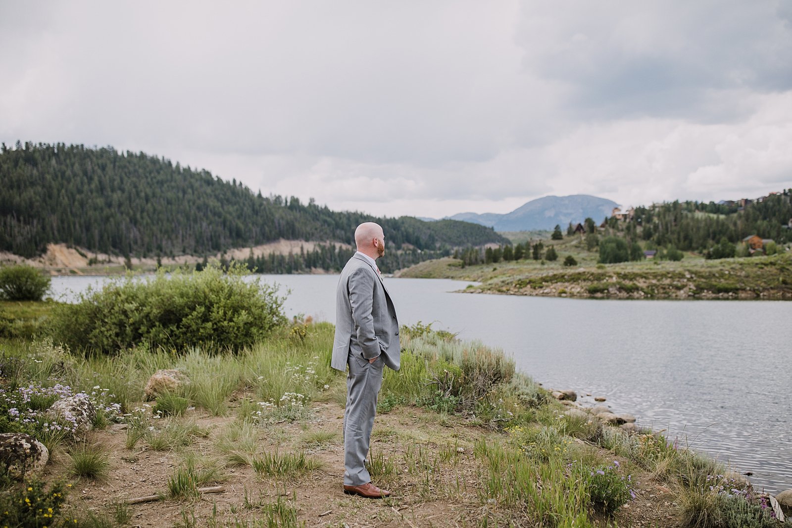 groom waiting on shoreline of lake for first look, groom in gray suit, dillon lake reservoir wedding, summit cove wedding, keystone colorado wedding, keystone colorado wedding photographer