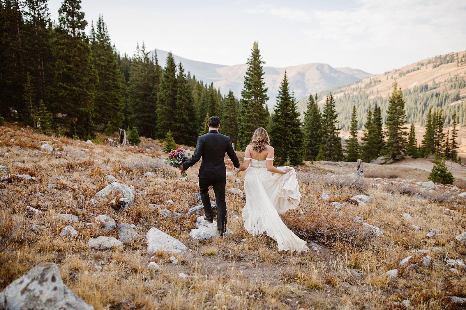 wedding couple hiking together, groom holding bridal bouquet, colorado trail elopement, gold hill elopement, peaks trail elopement, off the shoulder wedding dress, hiking wedding dress, mountain dress