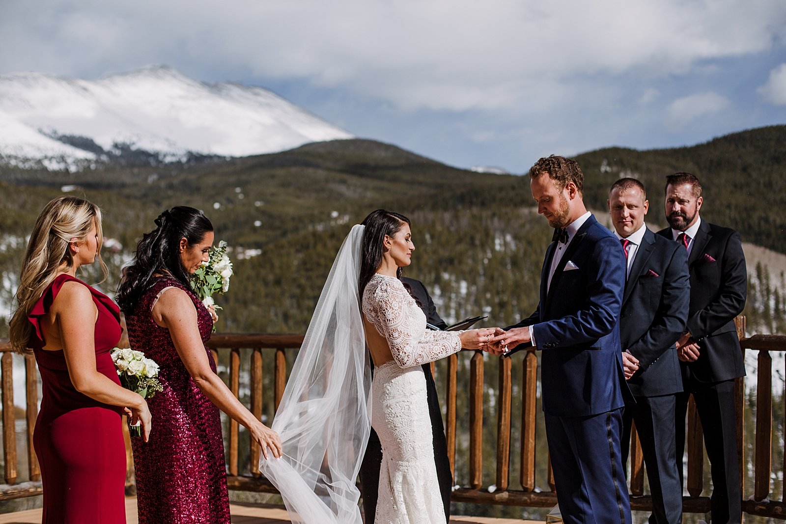 couple exchanging rings, the lodge at breckenridge wedding, the lodge at breckenridge ceremony, the lodge at breckenridge wedding deck, daughters of simone wedding dress, red bridesmaid dress