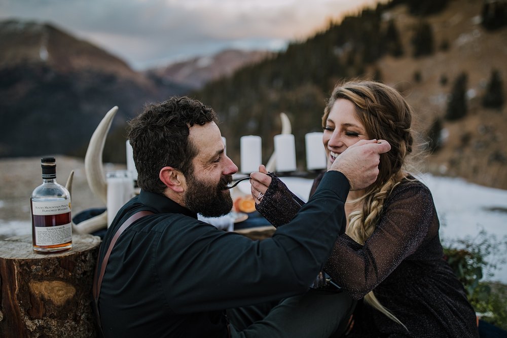 couple feeding one another wedding cake, spooky themed wedding cake, post elopement picnic, witchy wedding table settings, groom wearing all black, dark grooms attire, twilight mountain elopement