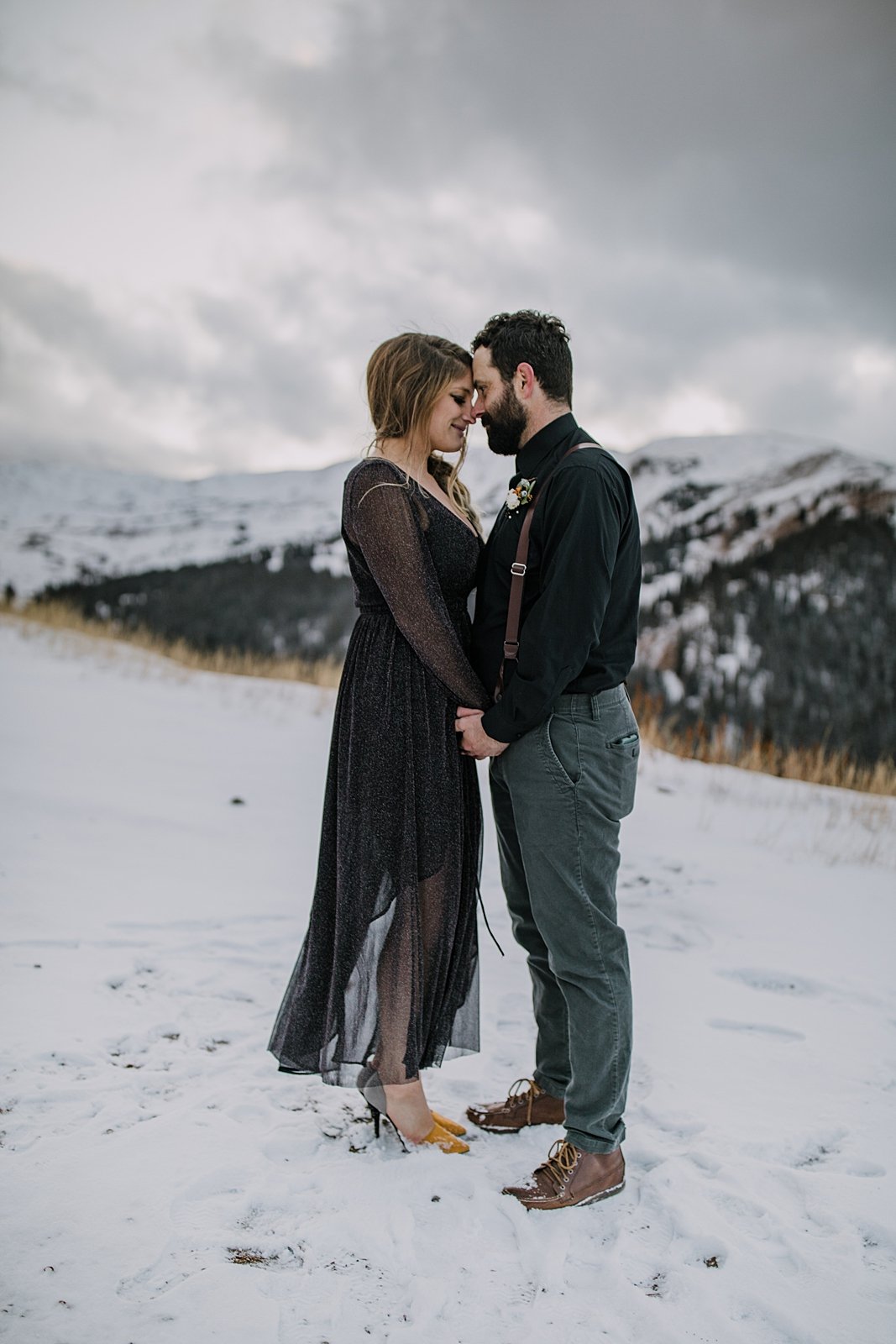 bride and groom holding each other close in the snow, black bridal attire, stormy mountain elopement, celestial inspired mountain pass elopement, dark wedding dress, snowy fall elopement