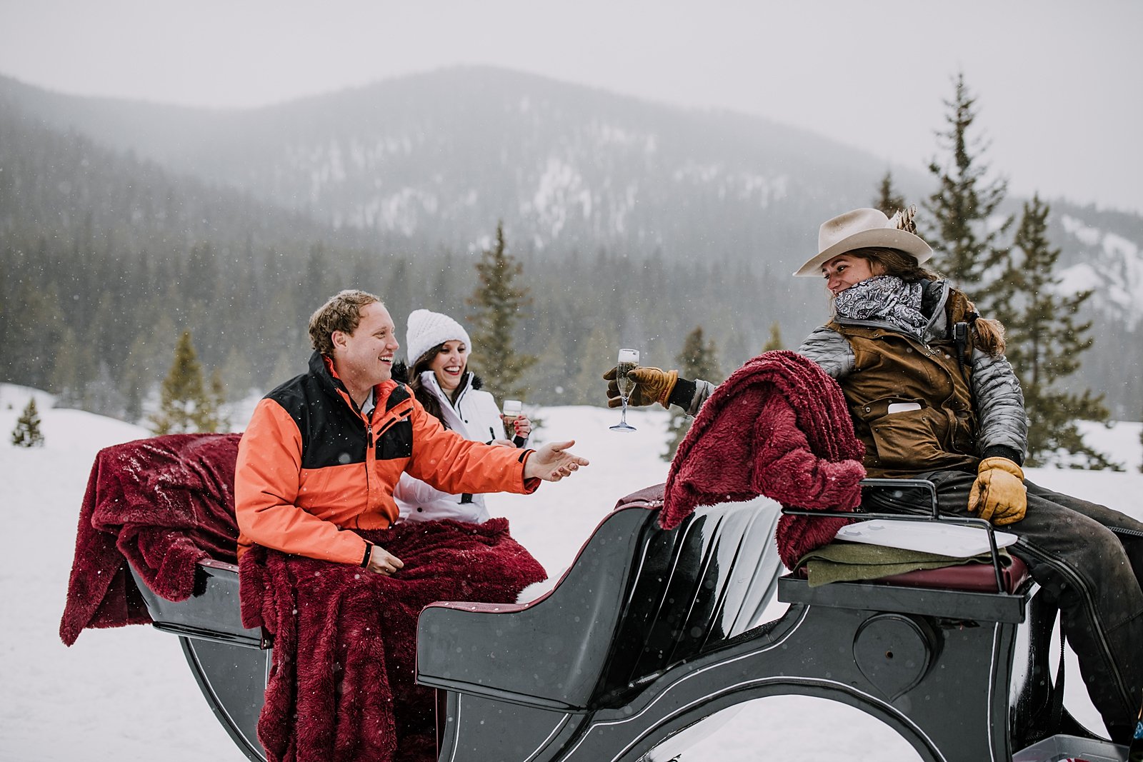 guide in hat and winter cowboy attire pouring champagne for couple while sitting on the sleigh, open one horse sleigh gliding through the snow, breckenridge spring break proposal, white bridal beanie