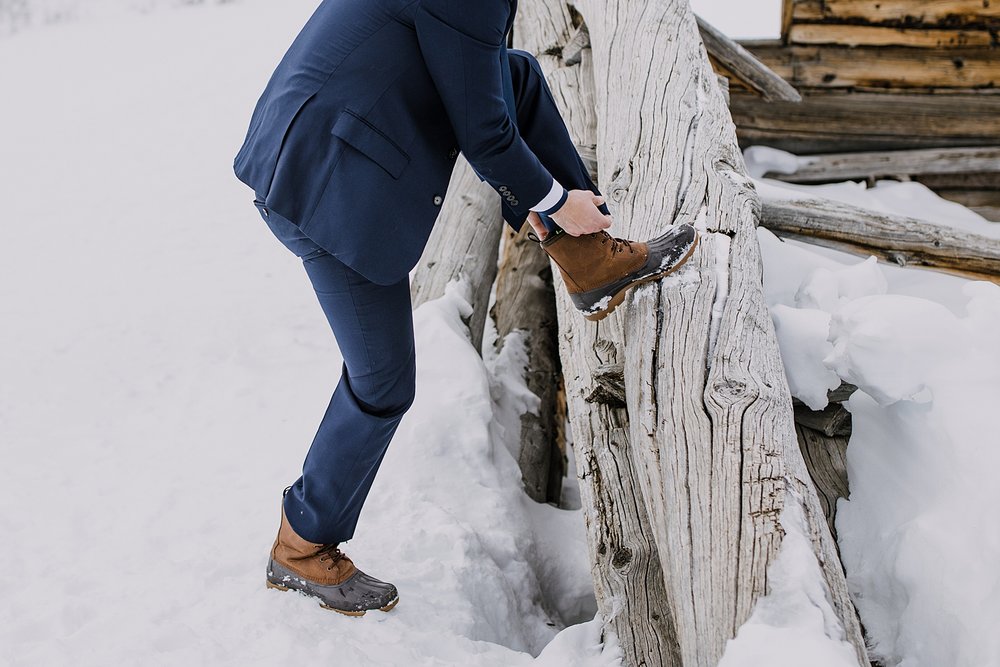 groom putting on hiking boots, boston mine cabins, hiking in the snow, colorado winter hiking, sorrel mens boots, custom wedding suit, leadville colorado, hiking wedding, elopement in leadville