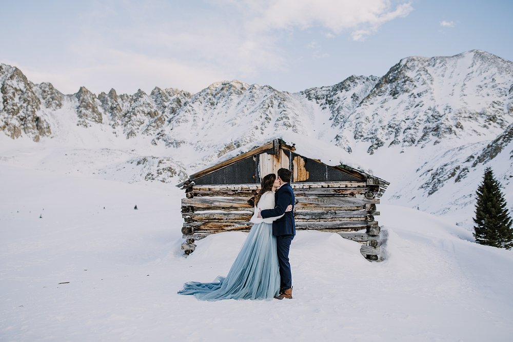 Bride kissing grooms cheek in front of log cabin, kiss in front of mining cabin, kiss in ghost town, leadville mining history, snowshoe to atlantic peak, kiss in front of quandary