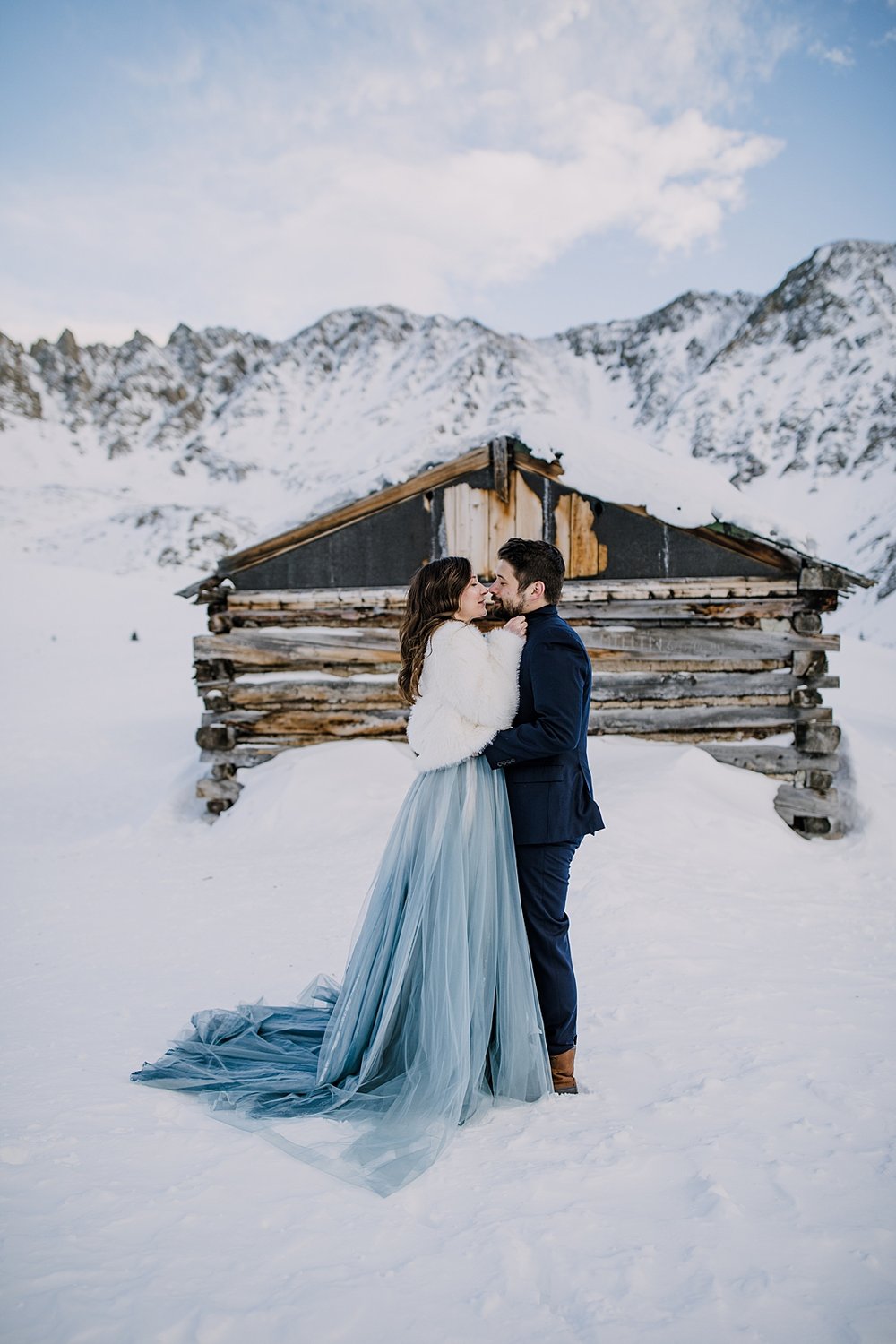 Couple snuggling in front of cabin, bride and groom snuggle in the mountains, snowy snowshoe adventure, snowy snowshoe elopement, kiss in front of cabin, winter wedding bridal jacket