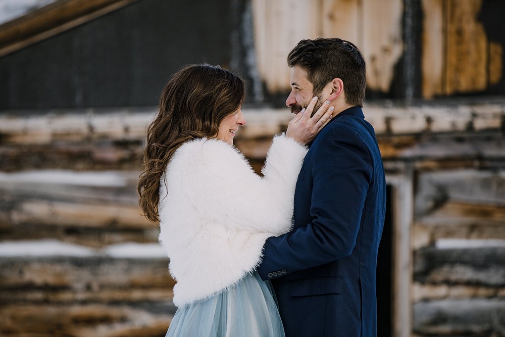 Bride warming hands on grooms face, bride and groom snuggle in the mountains, snowy snowshoe adventure, snowy snowshoe elopement, kiss in front of cabin