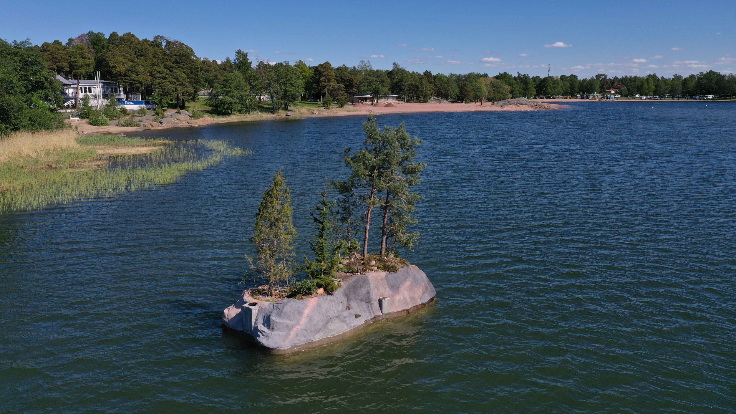 Overview of Raimo Saarinen’s Floating Island, a hand-sculpted concrete structure with living trees and coastal vegetation drifting on the sea in Rauma, Finland.