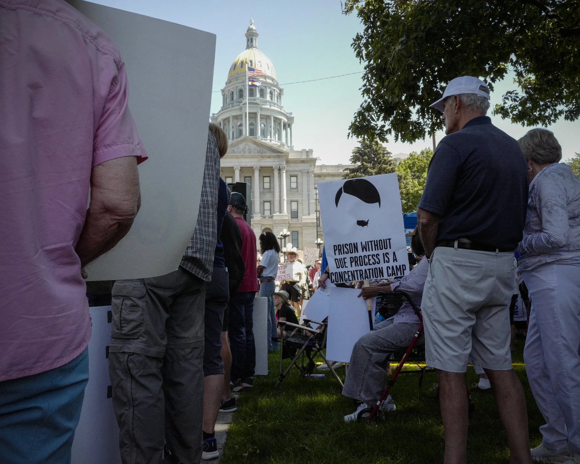Protesters gather on the lawn of the Colorado State Capitol building in Denver, holding a sign that reads, "Prison without due process is a concentration camp."