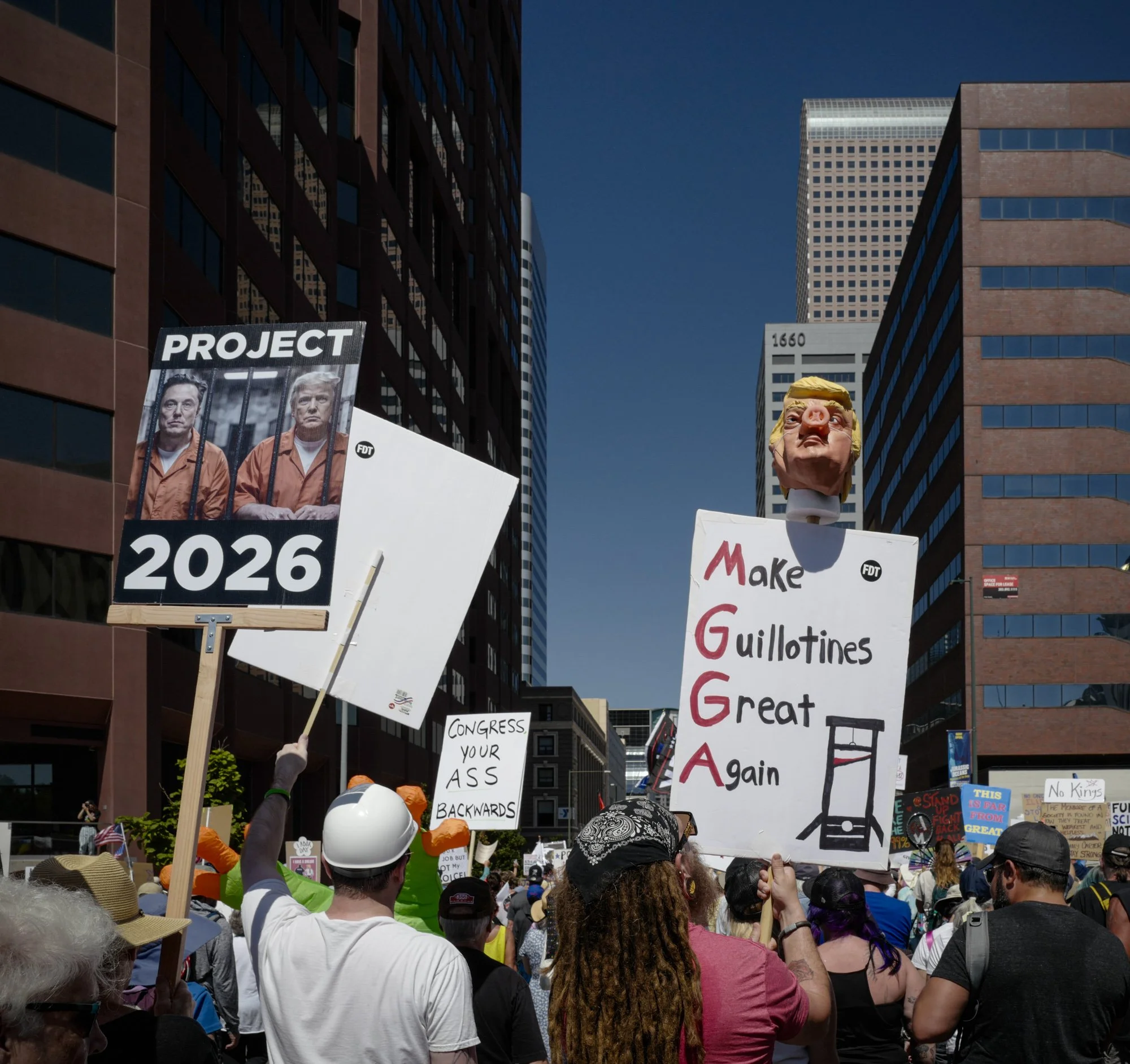 Protesters march through downtown Denver, carrying signs criticizing well-known figures. One sign shows a caricature of Donald Trump and Elon Musk in prison jumpsuits with the words "PROJECT 2026," while another states "Make Guillotines Great Again.
