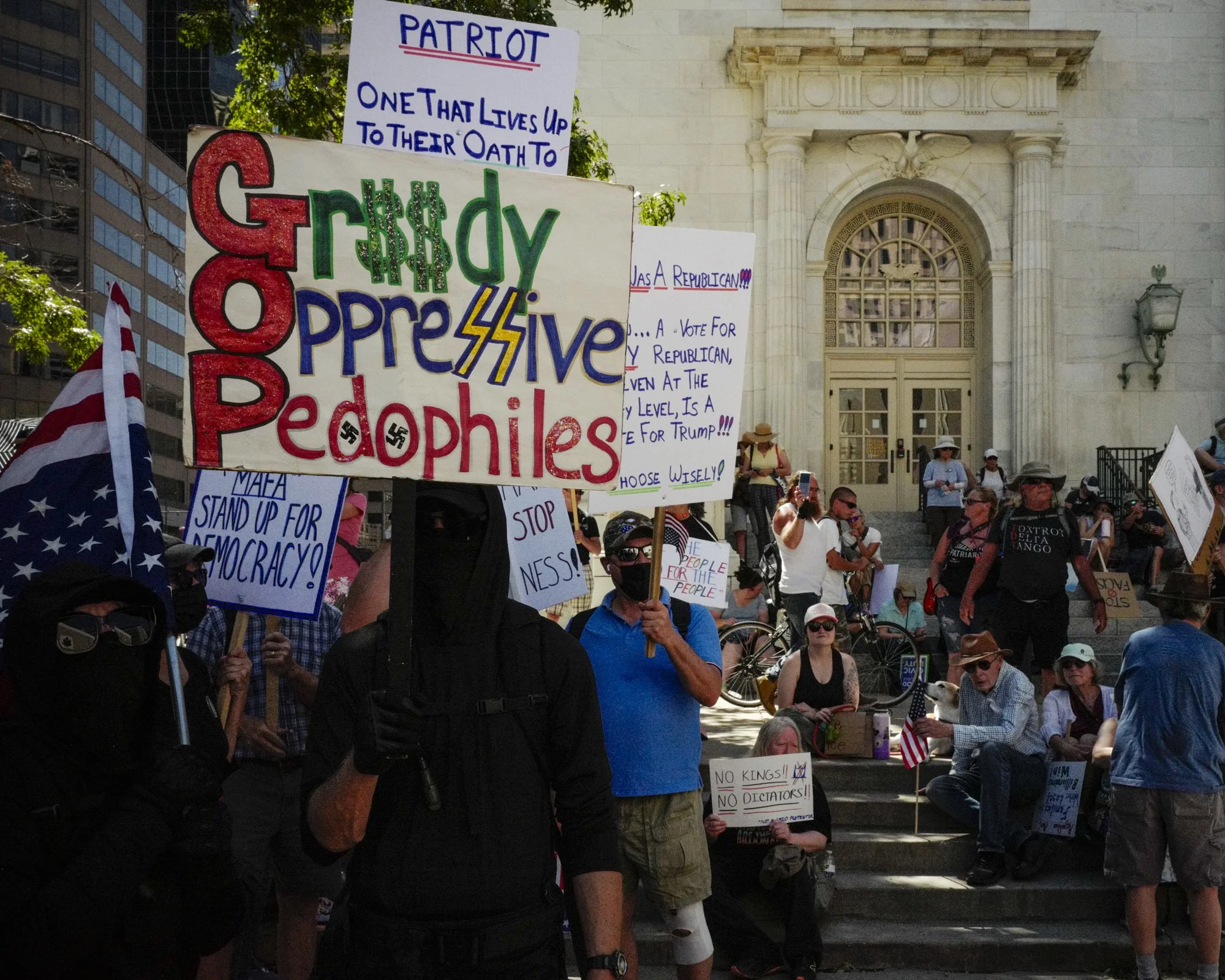 Protesters stand on a staircase outside a building in Denver. One person in dark clothing and a mask holds a sign with a prominent swastika symbol and the words "Greedy Oppressive Pedophiles."