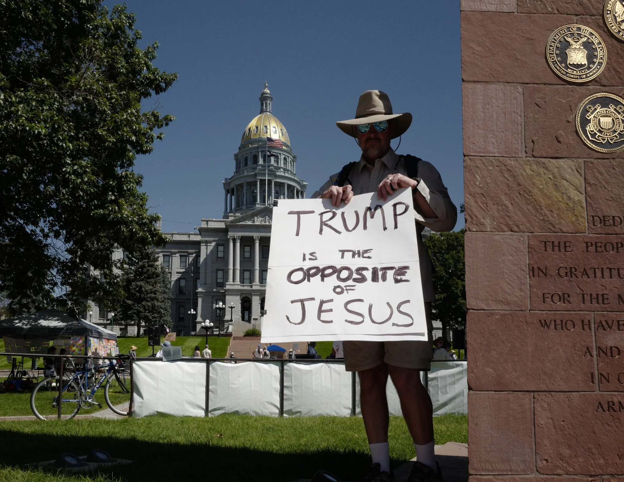 A person wearing a hat and sunglasses is holding a sign that says "TRUMP IS THE OPPOSITE OF JESUS," with the Colorado State Capitol building visible in the background.