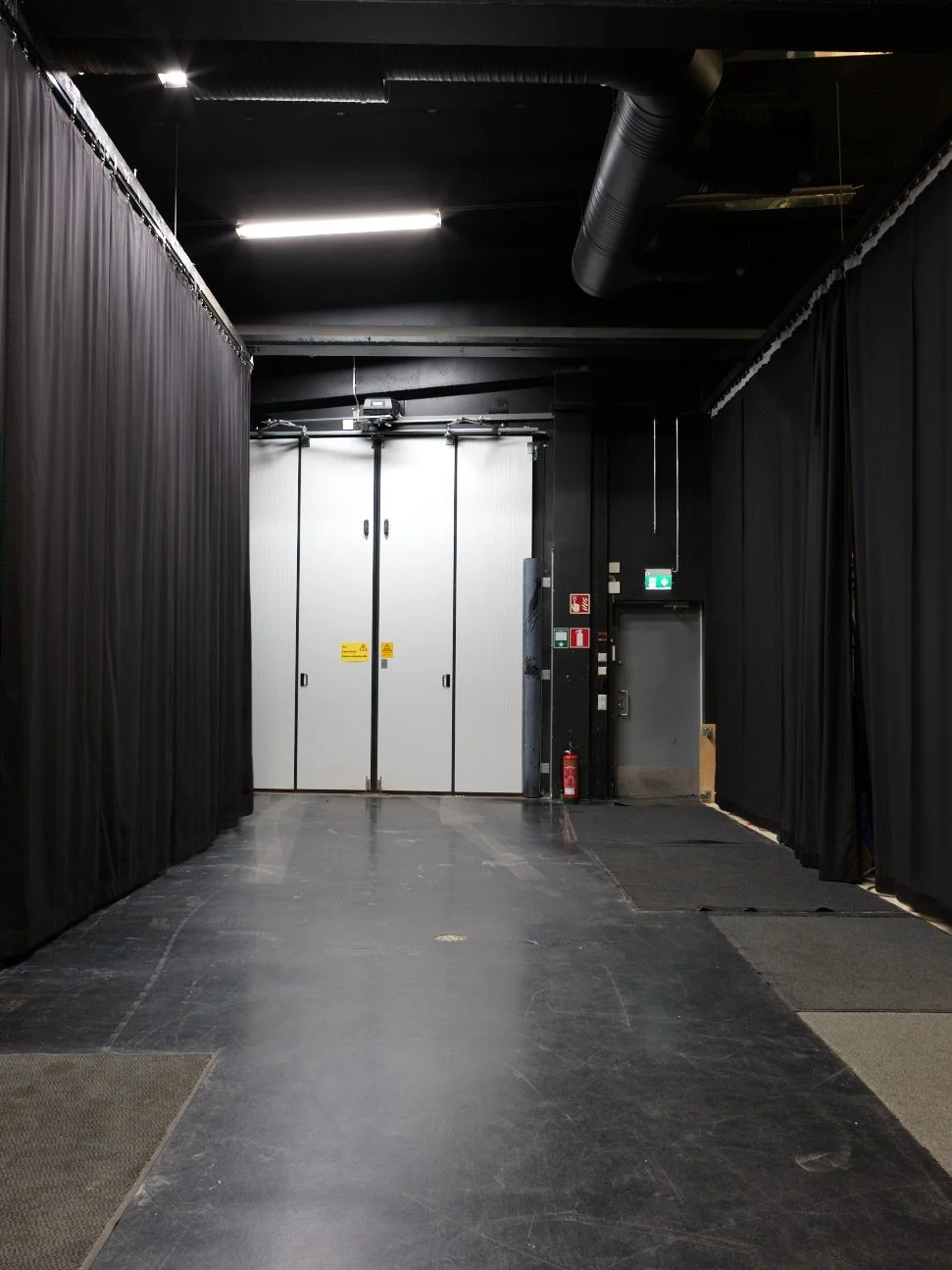 An empty backstage corridor with black curtains on both sides, leading to a white double door at the end, with emergency exit signs, fire extinguisher, and safety equipment visible.
