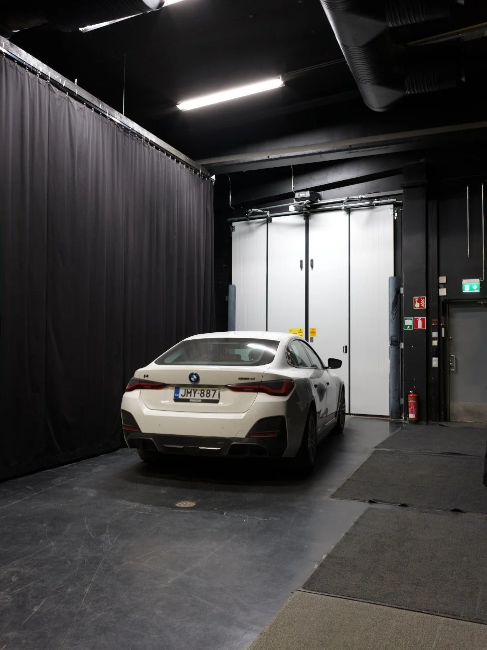 White BMW i4 parked inside a dark, industrial-looking garage with black walls, ceiling, and curtains, and metal double doors in the background.