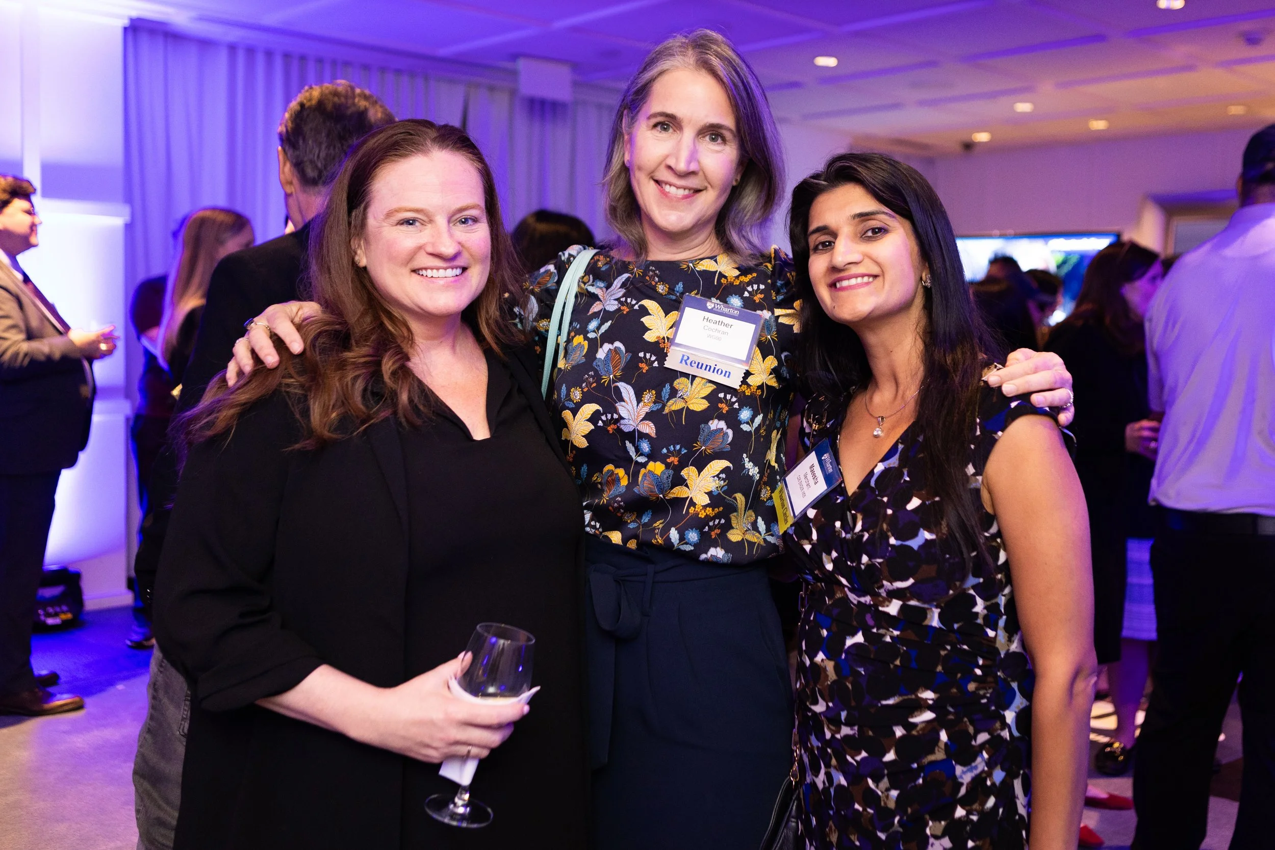 Three women smiling and posing together at a networking event, with a crowded background of other attendees and blue and purple lighting.