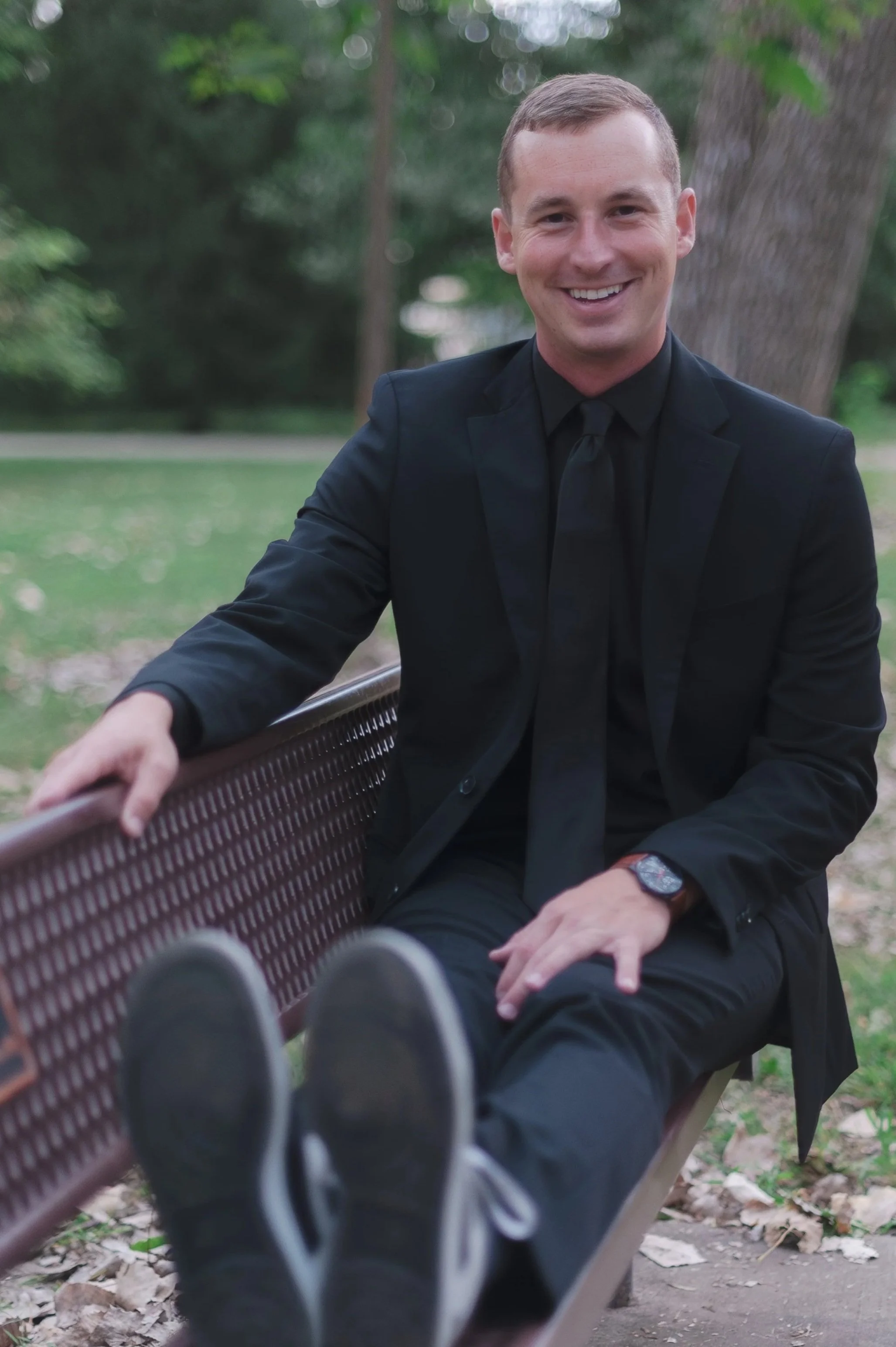 A man in a black suit and black shirt sitting on a park bench, smiling, with trees and green foliage in the background.