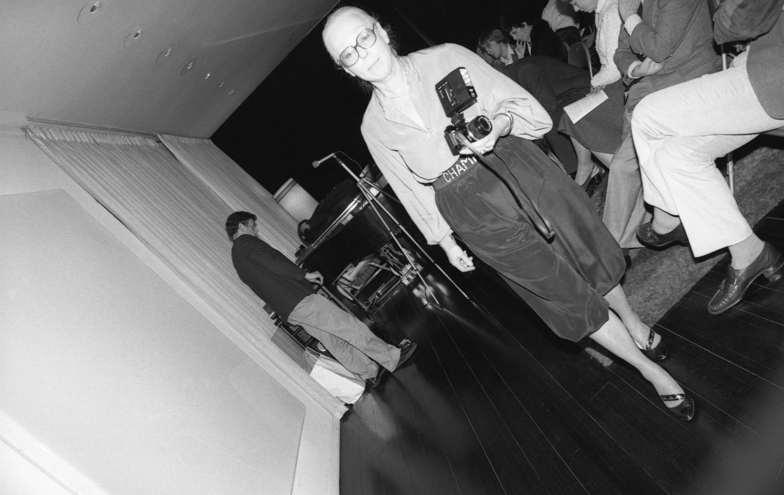 Betty Freeman with camera, Philip Glass at left in background. Betty Freeman's salon for Philip Glass at her house, Los Angeles, about 1981.