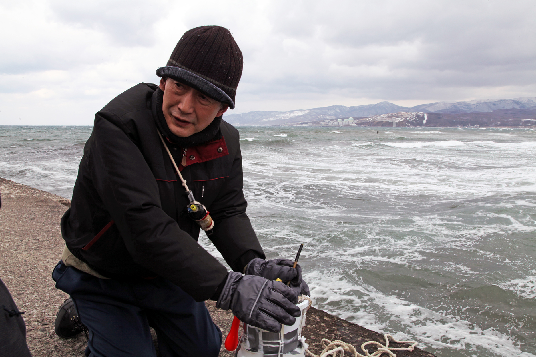 Takeichi Saito taking the temperature of the sea water at Iwanai. Tomari Nuclear Power Station is in the background. Image: authors.