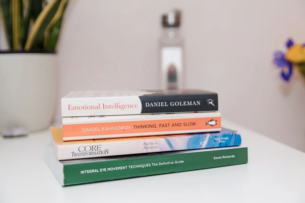 Stack of four books on a white surface with a plant in a white pot and flowers in the background.