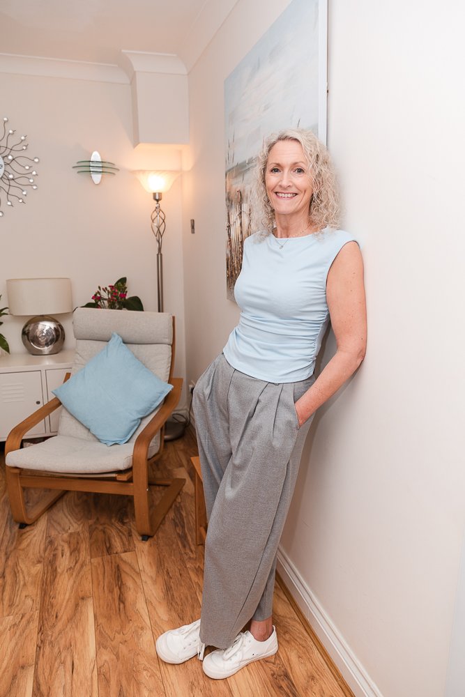 An older woman with curly gray hair smiling, standing in a cozy living room, leaning against a white wall, wearing a light blue sleeveless top, gray trousers, and white sneakers, with a painting and a chair with a blue pillow behind her.