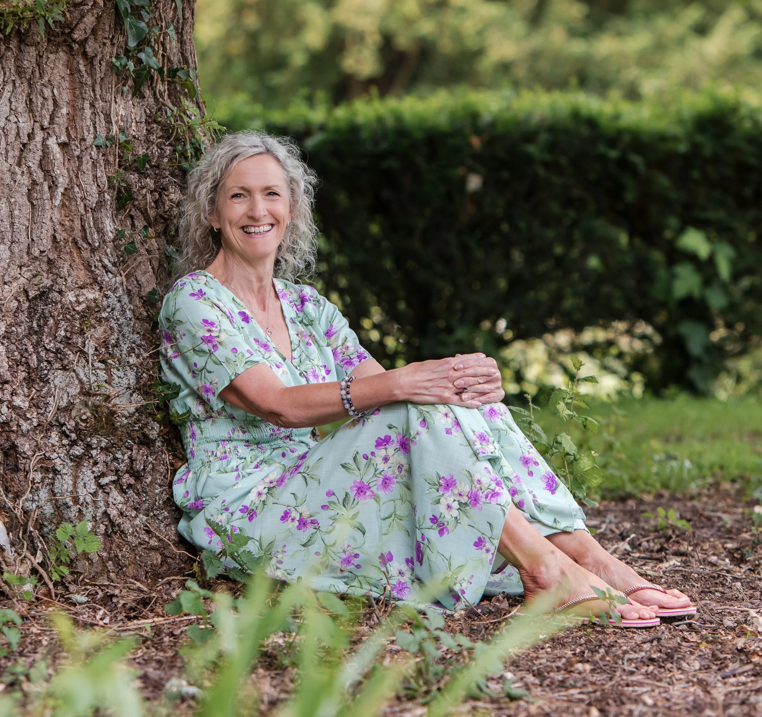 An woman with curly blonde hair smiling while sitting under an oak tree plants and flowers.