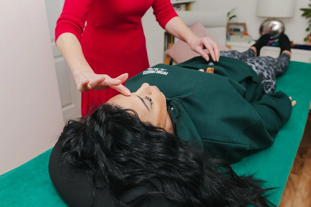 A woman lying on a massage or kinesiology table, receiving a treatment from a practitioner. The woman is on her back with her eyes closed, and the practitioner is gently touching her forehead. The room appears to be a cozy, home-like setting.
