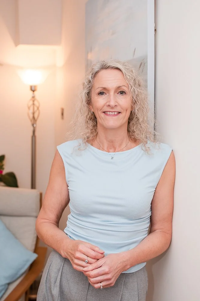 A smiling woman with curly blond hair standing indoors near a white wall, wearing a light blue sleeveless top and gray pants.