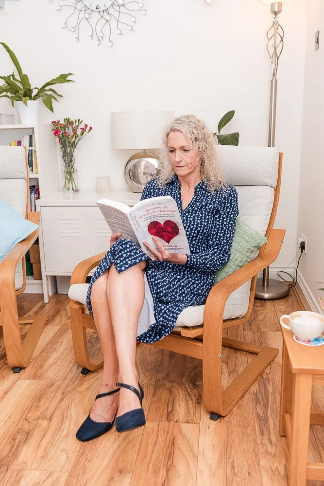 A woman with curly gray hair wearing a blue dress with white patterns and navy blue heels sits in a wooden armchair, reading a book with a red heart on the cover in a cozy, well-lit living room with wooden floors and decor plants.