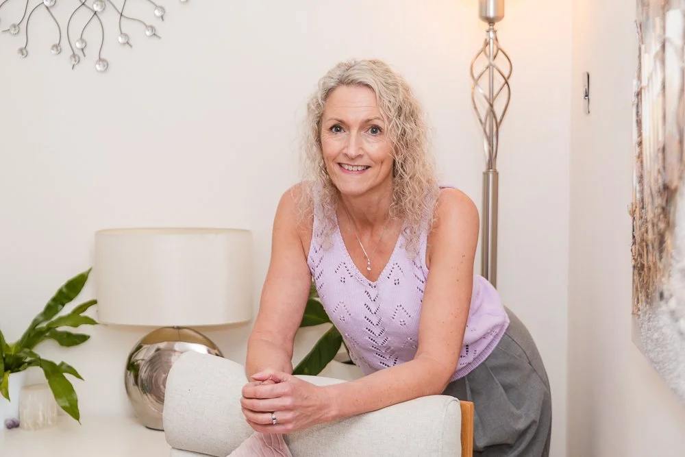 A woman with curly blonde hair smiling and leaning on a chair in a well-lit living room.