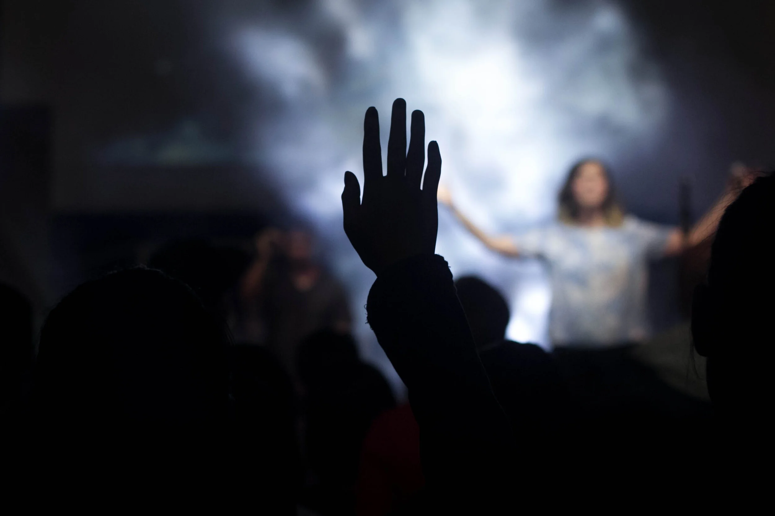 Silhouette of a person's hand raised in front of a blurred stage with a woman speaking or performing, and others in the background, with smoke or fog effects.