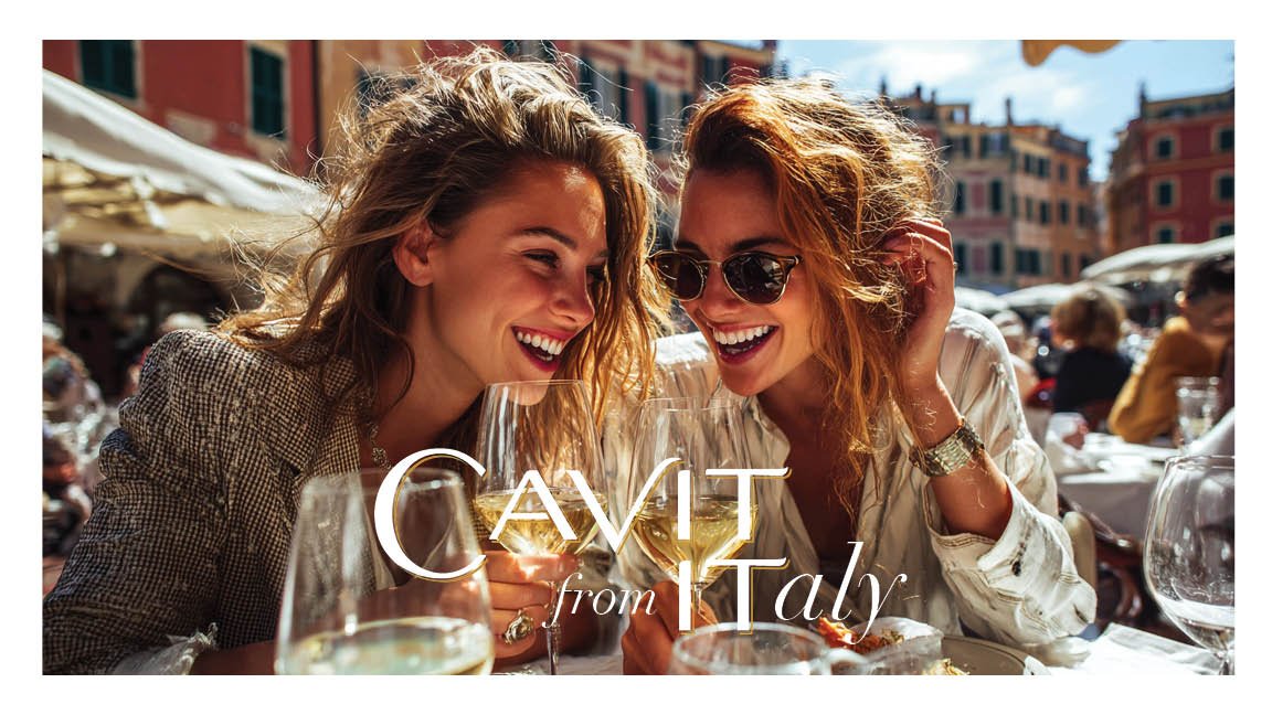 Two women enjoying drinks and smiling at an outdoor restaurant in Italy, with colorful buildings in the background.