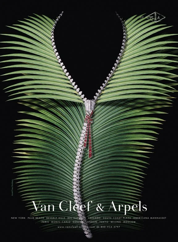 The image features a diamond necklace with a pendant, displayed on large green tropical palm leaves against a black background. The text at the bottom mentions Van Cleef & Arpels, along with store locations and website information.