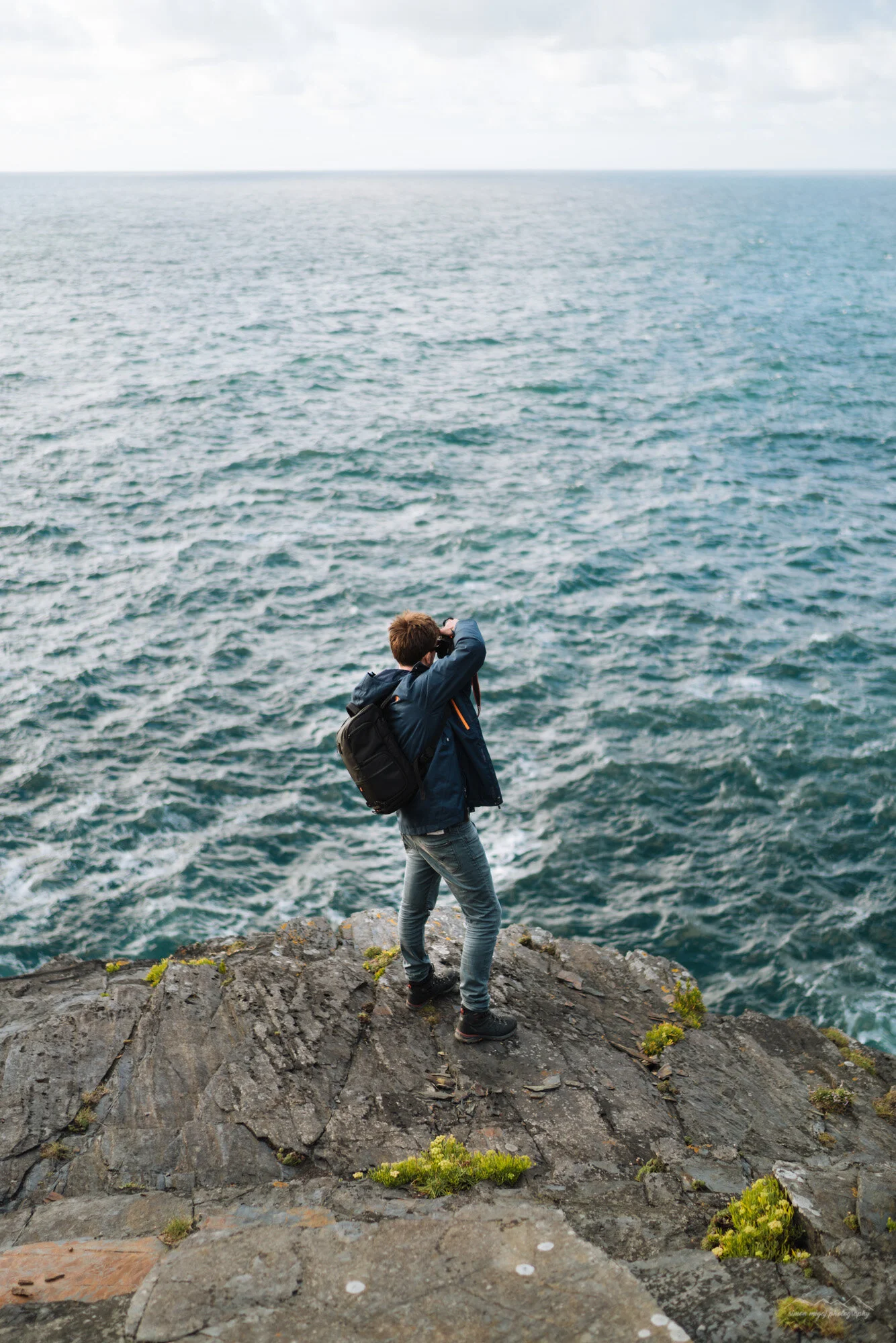 Young person standing on rocky edge overlooking the ocean, taking a photo, wearing a blue jacket, jeans, and a backpack.