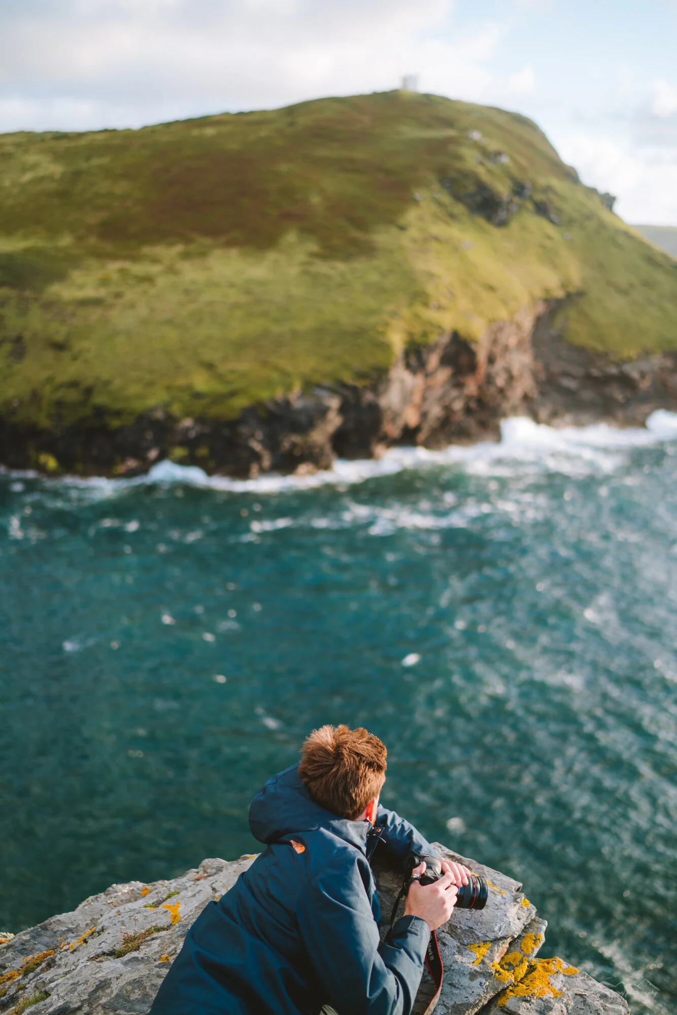 A person with brown hair wearing a dark jacket, sitting on a rocky shoreline, holding a camera, with a view of a large green island with cliffs in the background, and the ocean in the foreground.