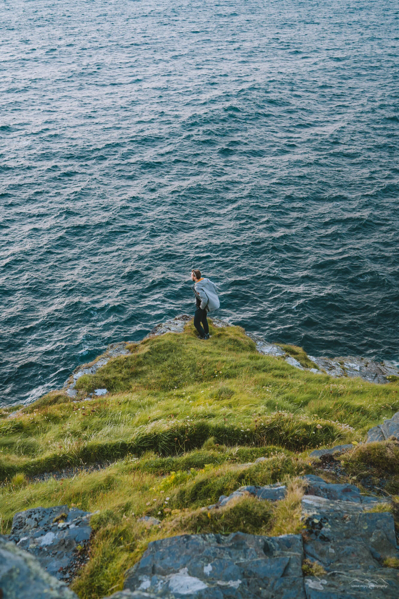 A person standing on a grassy, rocky cliff edge overlooking a large body of water.