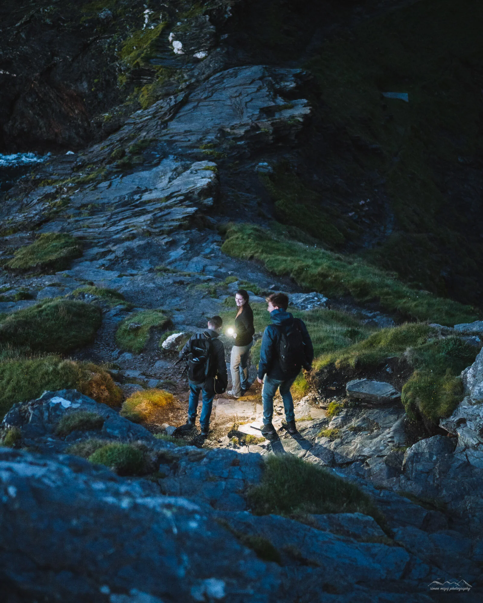 Four hikers walking along a rocky trail near a cliffside at dusk, illuminated by a headlamp, surrounded by green moss and rocks.