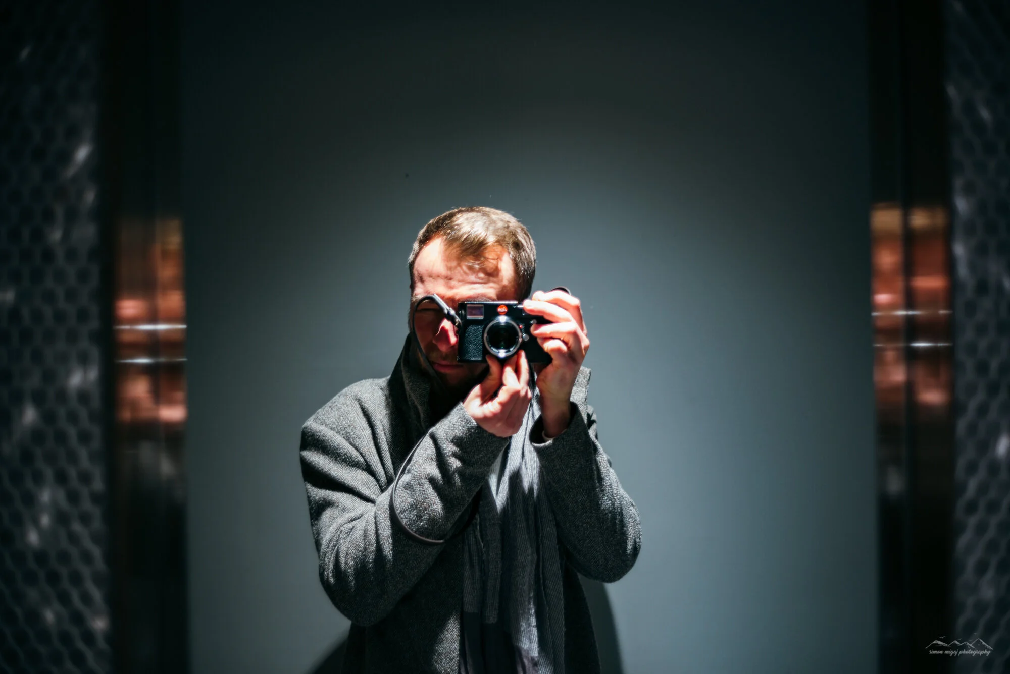 Man taking a mirror selfie with a camera in an elevator or enclosed space. The man is wearing a gray coat, and the background is plain with dark textured elevator doors on sides.
