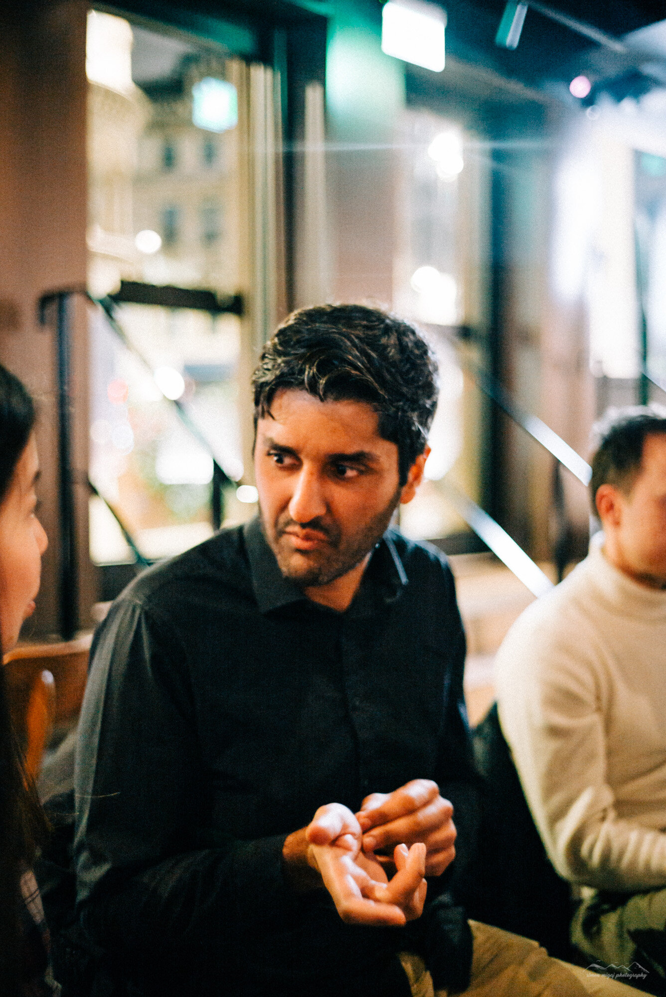 A man with dark hair and a beard wearing a black shirt, sitting in a restaurant or cafe, engaged in conversation with others. The background shows large windows with blurred cityscape outside, and warm lighting inside.