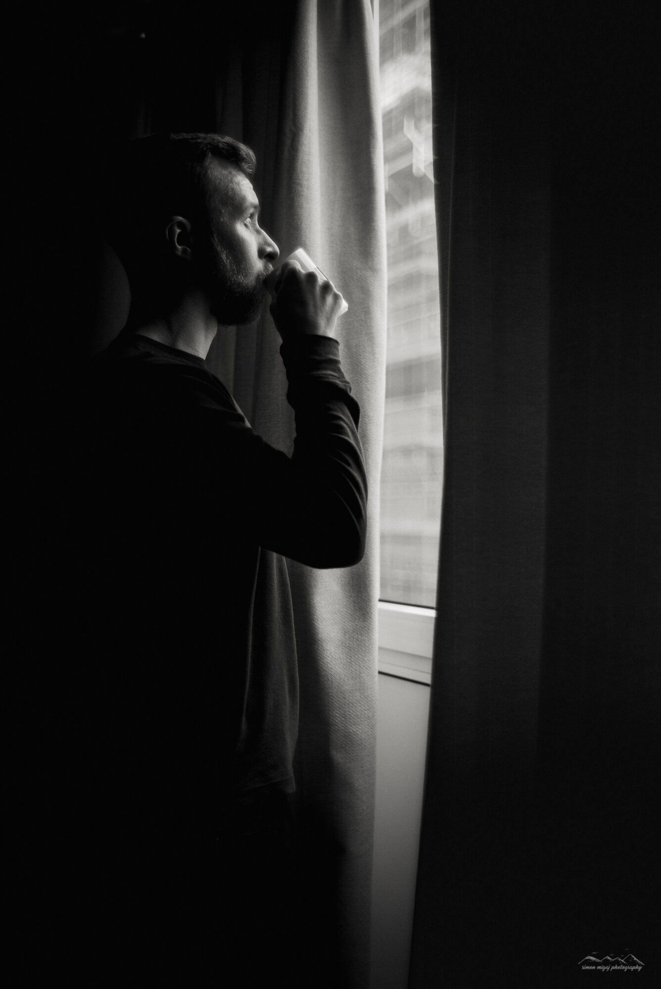 A black and white photo of a man with a beard looking out a window while holding a mug, with curtains and a building visible outside.