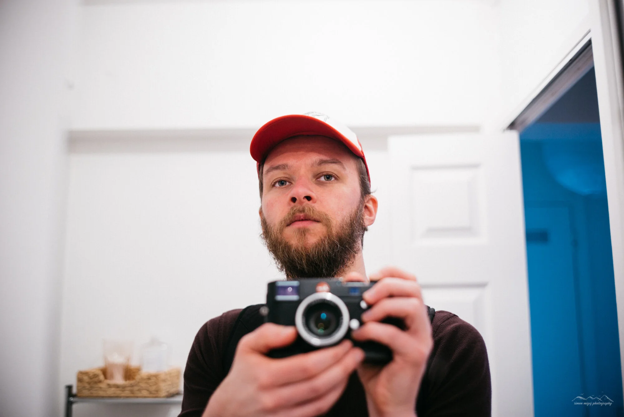 A man with a beard and mustache taking a selfie in front of a mirror using a black camera. He is wearing a red and white cap and a dark-colored shirt. The background includes a white wall, a wicker basket, and a shelf.