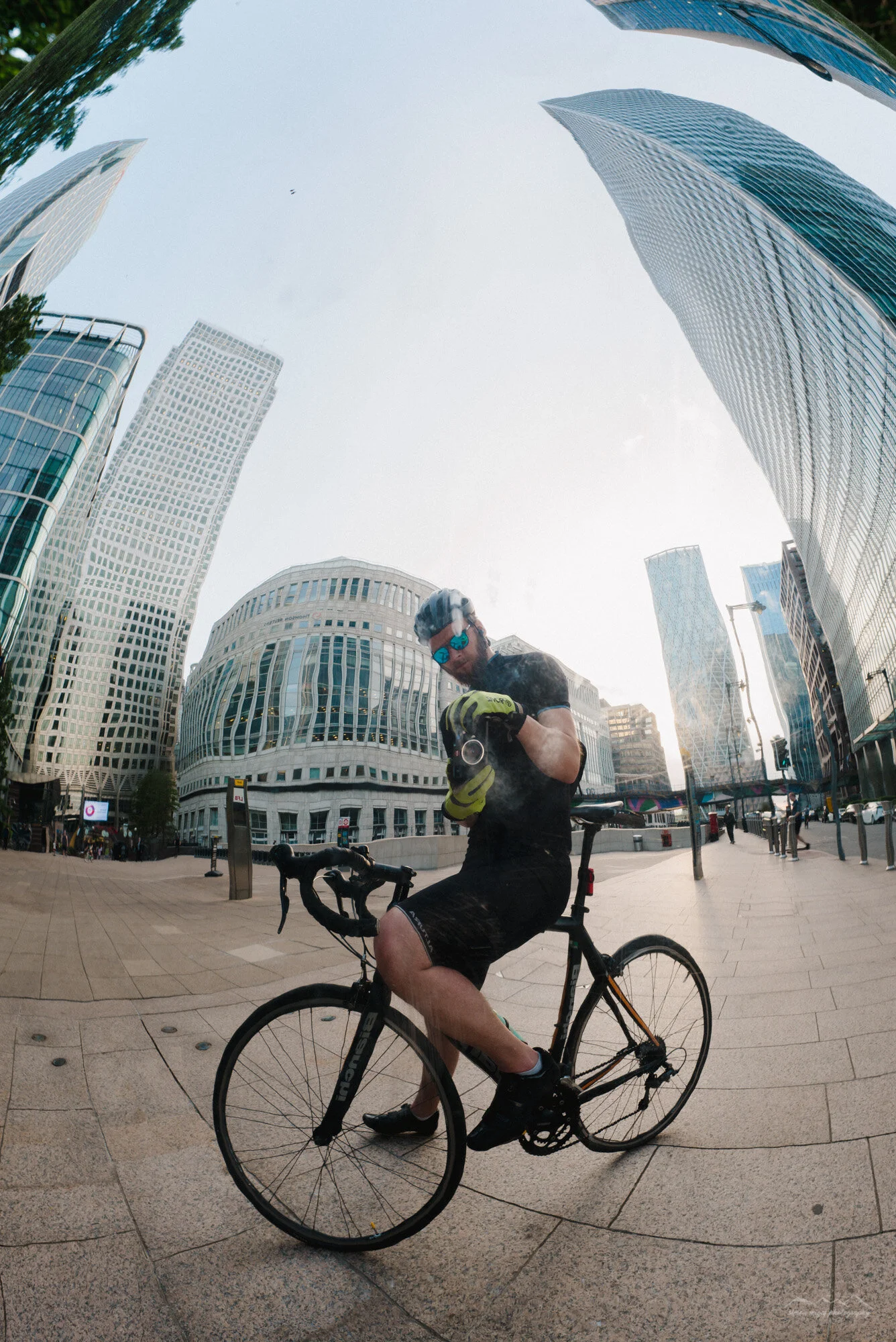 A man in cycling gear, sunglasses, and a helmet is sitting on a bicycle, adjusting his gloves, in an urban setting with tall office buildings and a clear sky.