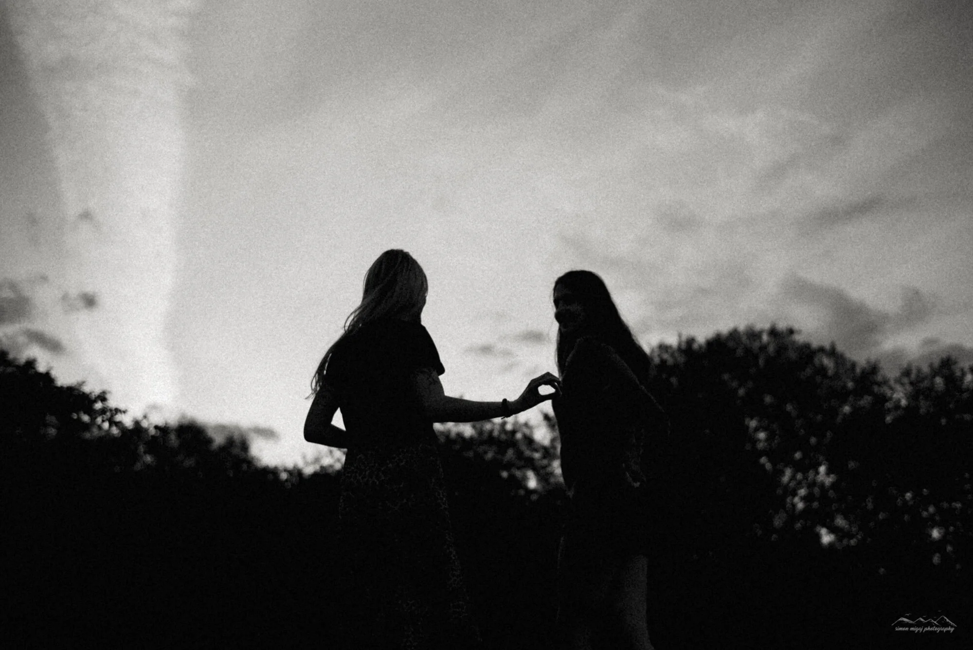 Silhouettes of two women holding hands outdoors during sunset, with a cloudy sky and trees in the background, in black and white.