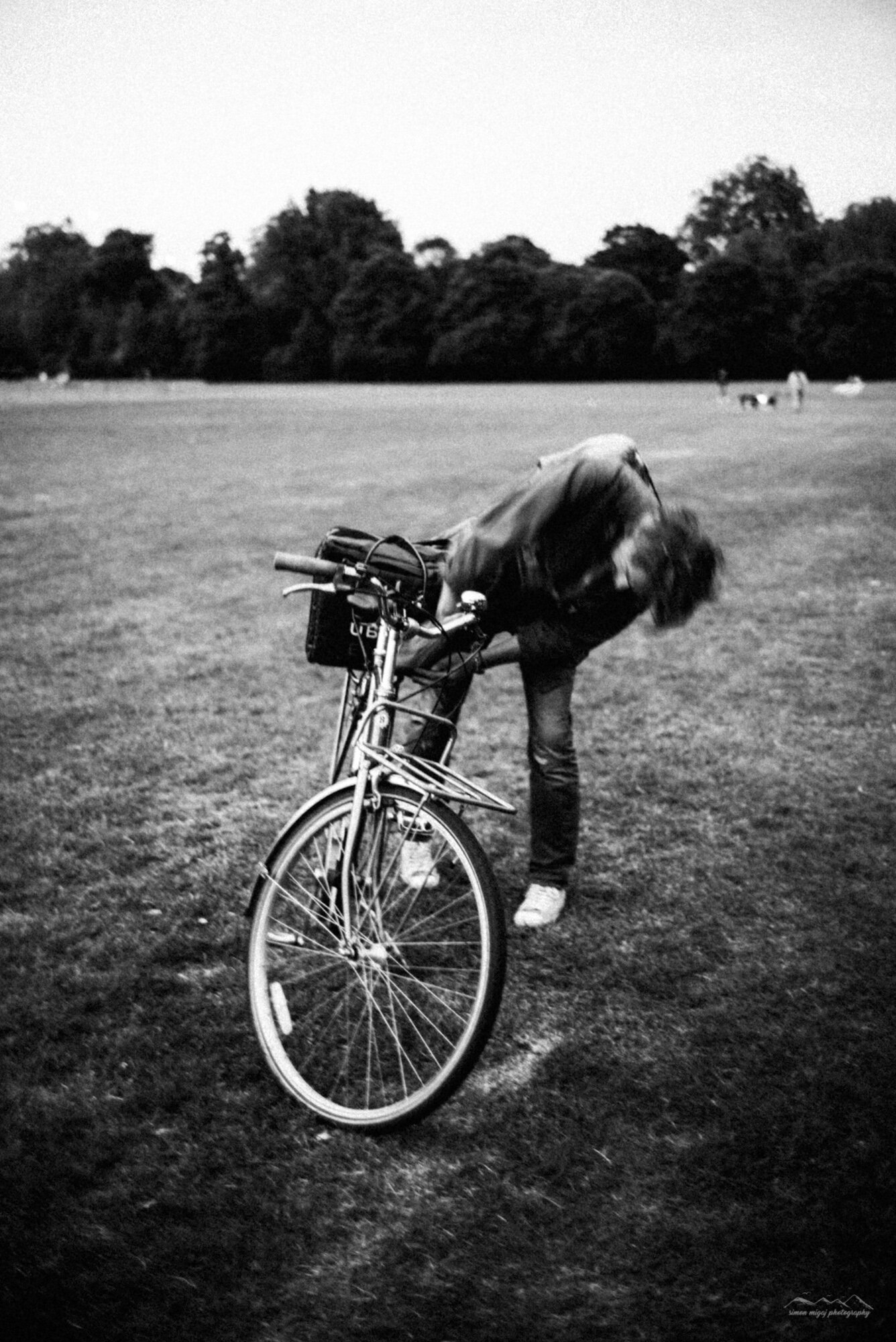 A person with long hair is bending over to hold a bicycle on a grassy field, with trees in the background. The image is in black and white.