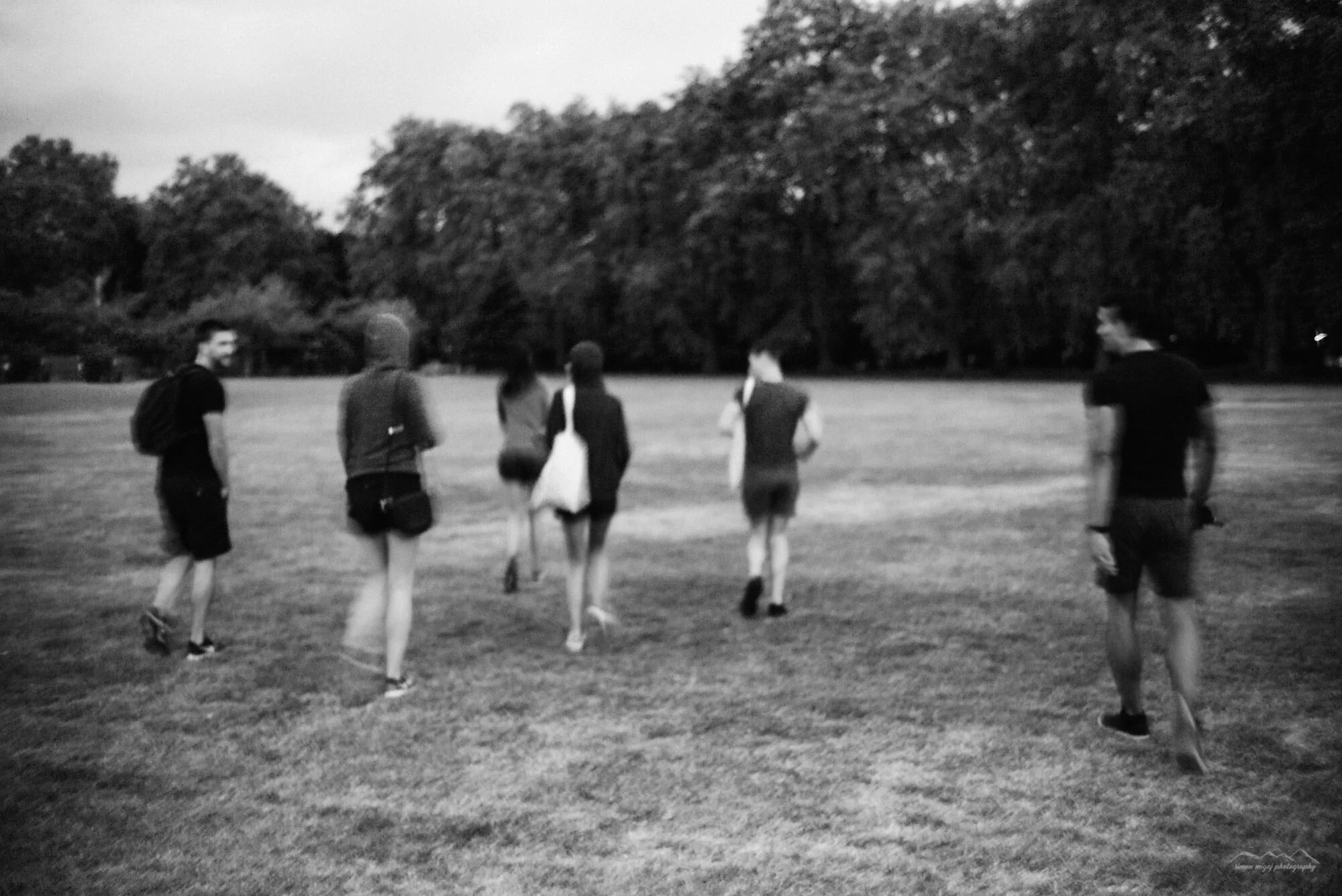 A group of young people walking across a grassy field in a park with trees in the background, black and white photograph.