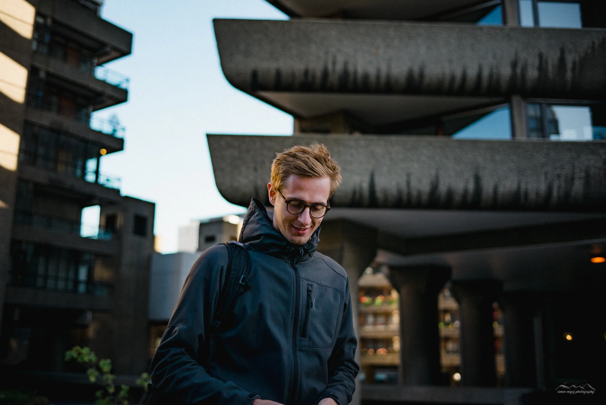 A young man wearing glasses and a navy blue jacket standing outdoors in front of modern apartment buildings with balconies, during evening or early night.