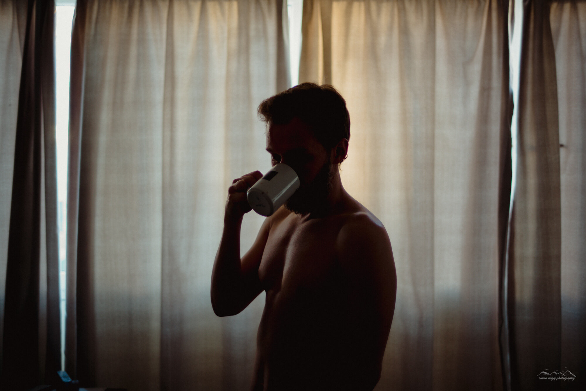 A shirtless man with a beard drinking from a mug backlit by natural light coming through curtains.