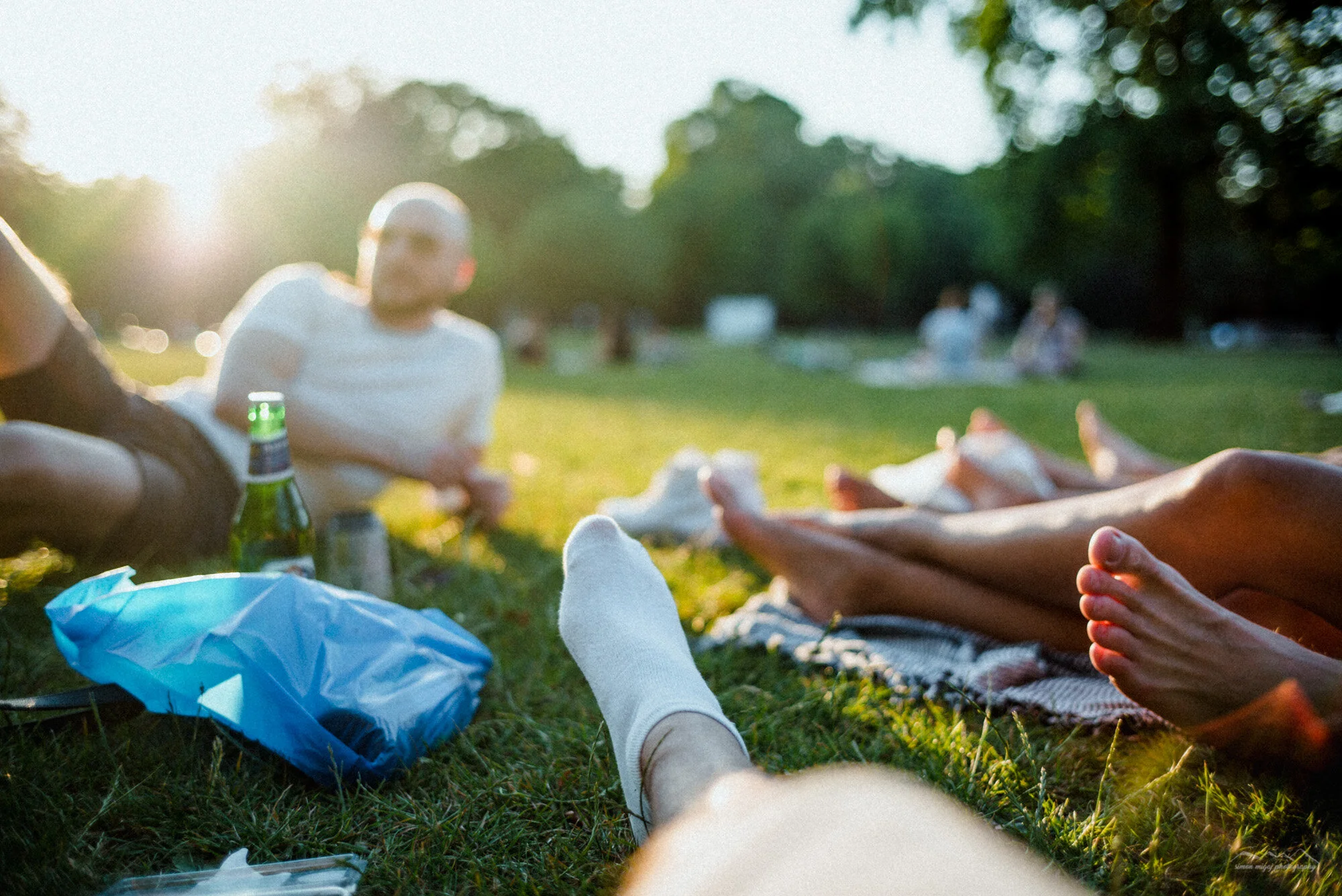 People relaxing on grass at a park during sunset, with others sitting in the background, some wearing white shirts, and a person with a bandaged foot in the foreground.
