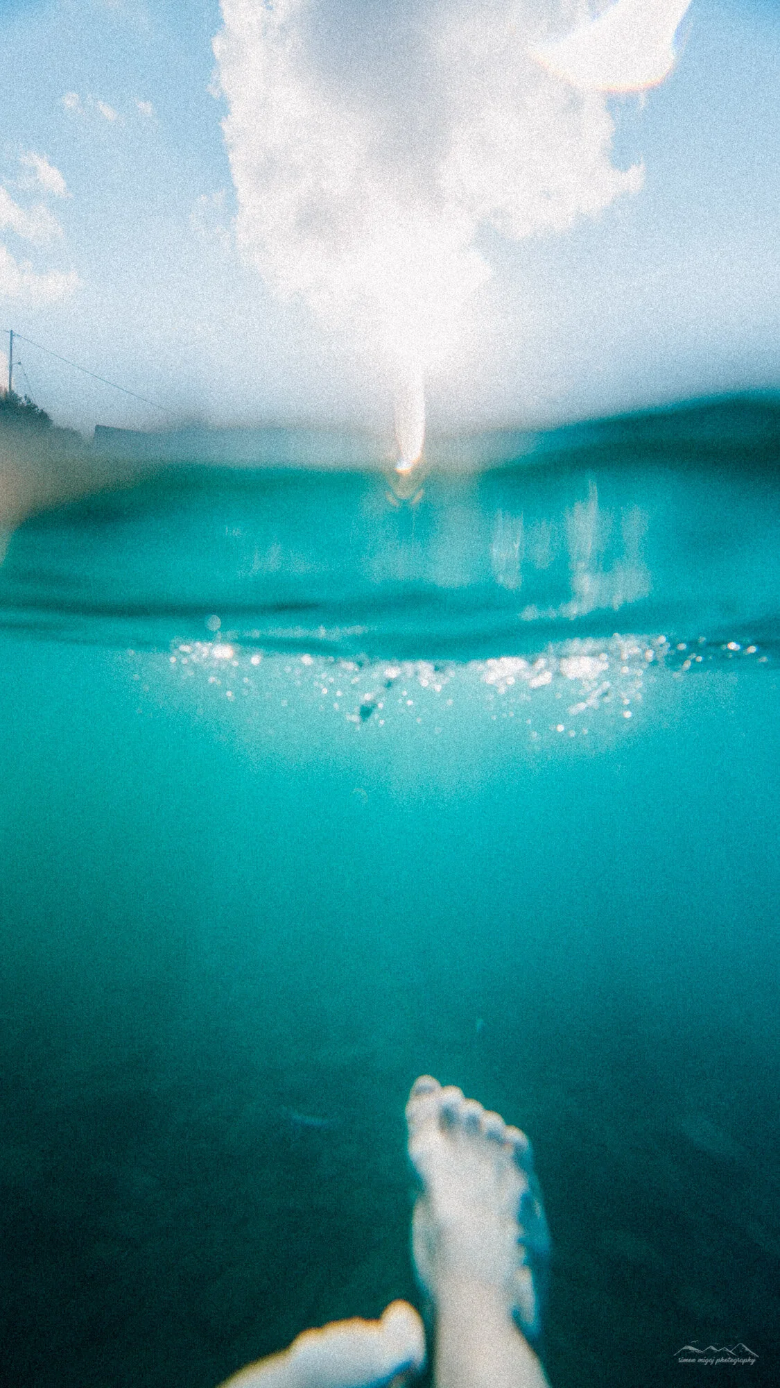 Underwater view of a person jumping into the water, with the photo split showing underwater and above water scenes, a cloudy sky, and a distant shoreline.