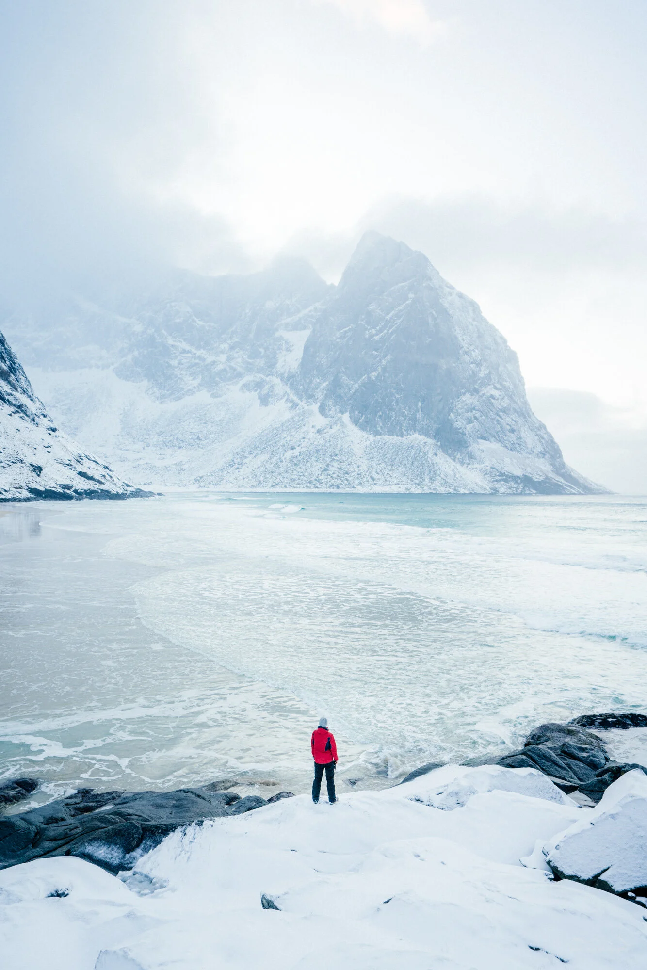 A person in a red jacket standing on snow-covered rocks and looking out over a partially frozen lake with snow-capped mountains and a cloudy sky in the background.